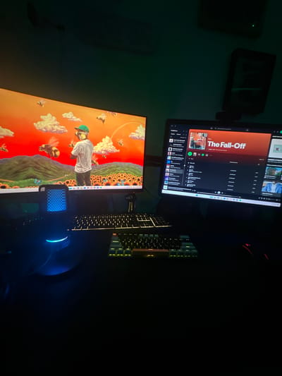 The image shows a desk with two computer monitors. The upper monitor displays Spotify playing "The Fall-Off." The lower monitor displays a colorful image with a person and bees. A keyboard and mouse are also on the desk. The mouse glows blue. The environment is dark. There is no information to suggest this is in Mérida, Mexico.