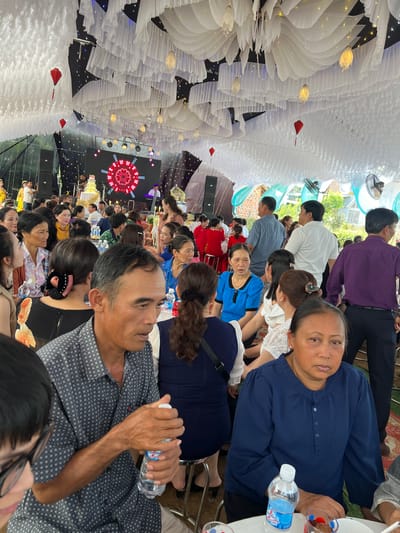 This image captures a vibrant indoor celebration in Bình An, Vietnam, featuring numerous attendees gathered for an event, likely a wedding or a large family reception. The scene is bustling with people of varying ages, predominantly adults, seated at tables arranged for dining.

The venue's ceiling is elaborately decorated with white pleated fabric, resembling layered clouds or petals, interspersed with small warm-toned string lights and larger, flower-shaped decorative lamps. Several red lanterns hang overhead, adding contrasting color. Ceiling fans are visible within the decorated structure.

Towards the back of the room, a stage area is discernible, equipped with large speaker boxes and a prominent circular screen displaying an intricate red and white geometric pattern. A multi-tiered cake is also present near the stage, further suggesting a celebratory occasion.

In the foreground, an older woman in a blue blouse looks directly towards the camera with a neutral expression. To her left, an older man in a patterned short-sleeved shirt holds a "Lavie" branded water bottle, his attention directed towards the stage area. Other guests are scattered throughout the venue, some engaged in conversation, while others observe the activities on stage. Tables are set with plates, cups, and additional "Lavie" water bottles. The overall ambiance appears relaxed and festive, indicating a significant social gathering taking place during what seems to be a well-lit daytime event.
