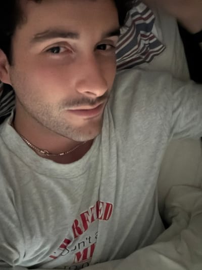 A selfie-style image depicts a male subject lying down, positioned in what appears to be a bed. The individual has short dark hair, dark eyes, and light facial stubble. He is wearing a light grey short-sleeved t-shirt, which features partially visible red lettering on the chest, including the word "ARMED" and what appears to be "don't MI" below it. A thin metallic chain necklace is worn around his neck, with a small, circular clasp visible at the front. Behind his head, a pillow with prominent red, white, and blue stripes is visible, along with light-colored bedding or sheets. The subject's gaze is directed towards the camera, and his mouth is closed, forming a slight, subtle curve. The immediate environment suggests an indoor setting, likely a bedroom. The image provides no discernible features, landmarks, or unique environmental details that would allow for precise geographical determination within the city of Santo Domingo, Chile.