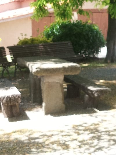 A stone picnic table with two stone benches sits in a park-like setting. The table is rectangular and appears to be made of rough-hewn stone blocks. One bench is visible to the left of the table, and another is partially visible to the right. A dark wooden bench is in the background behind the stone table. The scene is outdoors, with dappled sunlight suggesting it is daytime. A large, leafy tree is visible in the upper right portion of the image, casting shadows on the ground. The ground is covered with a mixture of grass and fallen leaves. In the background, there is a building with a reddish-tiled roof and a wall painted a light color. A dense green bush is situated behind the stone picnic table. The overall impression is a peaceful outdoor setting, likely a park or picnic area. There is no visible text in the image. The image appears to be taken in Susqueda, Spain.
