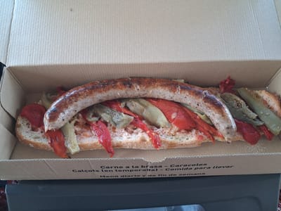 A close-up, high-angle shot shows a long, split bread roll filled with a grilled sausage, roasted red peppers, and what appear to be grilled leeks or asparagus. The food is presented in a brown cardboard takeaway box. The text printed on the side of the box is in Spanish and reads "Carne a la brasa - Caracoles Calçots (en temporada) - Comida para llevar Menú diario y de fin de semana". This indicates the location is likely a restaurant in or near Torrefarrera, Spain, serving grilled meats, snails, and calçots (a type of spring onion) as takeaway options, with daily and weekend menus. The lighting is natural and even, and the focus is sharp on the food.
