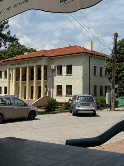 A daytime outdoor shot depicts the Narodno Chitalishte "Probuda 1941" cultural center in Kardam, Bulgaria. The building is a two-story structure with a red tile roof and beige exterior. The facade features a colonnaded entrance. Two silver cars are parked on the adjacent street. Utility poles and wires are visible. A sign is seen on the left.