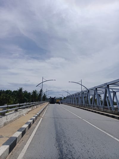 The image shows a perspective view of a grey steel truss bridge spanning over a body of water. The bridge has two lanes of asphalt roadway with a white center line and white edge lines. A sidewalk runs along the left side of the bridge, separated from the road by a concrete curb with alternating black and white painted sections. Metal railings line the edge of the sidewalk.

On the left side of the bridge, a dense line of palm trees and other tropical foliage is visible beyond the railings. On the right side, the steel structure of the bridge is prominent, with diagonal supports forming a geometric pattern.

Two lampposts with curved arms are present on both sides of the bridge, spaced along its length. In the distance, a black truck with a yellow cab is driving on the bridge. A person on a motorcycle is also visible, further ahead on the right lane.

The sky above is partly cloudy with white and grey clouds against a pale blue background.

The location is identified as the bridge in Neuboknaleung, Indonesia.