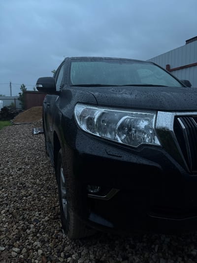 The image shows a black SUV parked on a gravel surface under an overcast sky. The vehicle's headlights and grille are visible, with water droplets on the hood and windshield, indicating recent rainfall. In the background, there is a pile of sand or soil, some stacked lumber, and a corrugated metal fence. The scene appears to be in a rural or semi-rural setting, consistent with the village of Shebarshino, Russia. There are no people or animals visible in the image.
