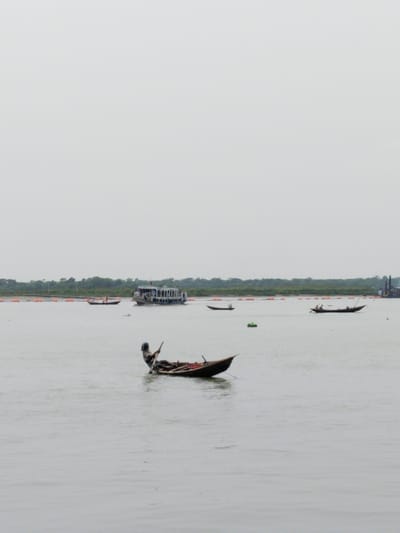 A man rows a wooden boat in a river in Pangasia, Bangladesh. There are two other small boats and a larger ferry in the background. The river is wide and calm.  The sky is overcast and there are trees visible on the far shore.  The scene is located at latitude: 22.704, longitude: 90.565.