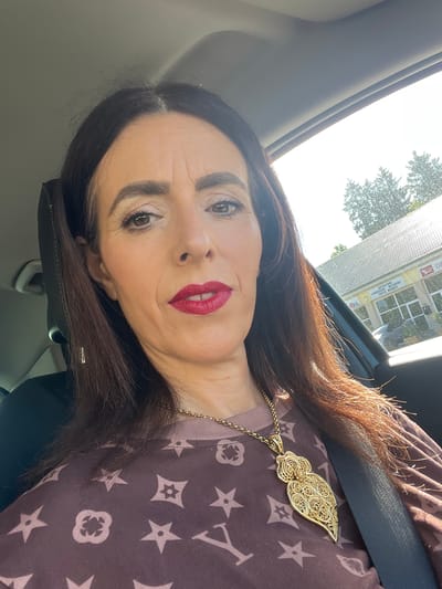 This is a selfie taken by a woman inside a car, likely in Roodt-sur-Syre, Luxembourg, given the signage visible in the background. The woman has dark brown hair, styled down with some flyaways. She is wearing makeup, including red lipstick and eyeshadow. She has a prominent gold necklace with an intricate, heart-shaped pendant. Her top is dark brown with a repeating pattern of intertwined symbols and stars. The interior of the car is visible, including the headrest and the seatbelt. Outside the car, through the window, a building with signage is visible, appearing to be a repair shop or garage with French text. The sky is bright and sunny, with trees visible behind the building.