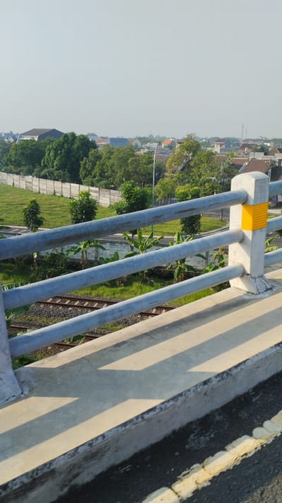 The image depicts an elevated view from a concrete bridge railing overlooking a suburban landscape in Trosobo, Indonesia. The railing, constructed of concrete posts and gray metal pipes, dominates the foreground, casting long shadows on the concrete walkway. Below the railing, railway tracks run horizontally across the scene, bordered by green grass and banana trees. In the middle ground, a concrete wall separates the railway area from a grassy field with scattered trees. Beyond this, a cluster of houses with red-tiled roofs and various building materials are visible, interspersed with more greenery and a distant street light. The sky is clear and a pale blue, suggesting a bright, sunny day. There are no people or animals visible in the frame.