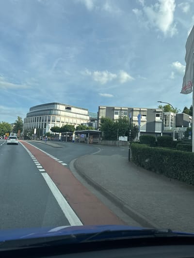 The image shows a street scene in Wetzlar, Germany, with a large, modern, curved building on the left and a smaller, rectangular building on the right. A white car is driving on the left side of the road, in the same direction as the camera. There is a red lane marker on the road, and a sidewalk with a hedgerow next to it. A white flag with red lettering is visible on the right side of the image, attached to a pole. The sky is cloudy. The scene appears to be a business district or an office park.