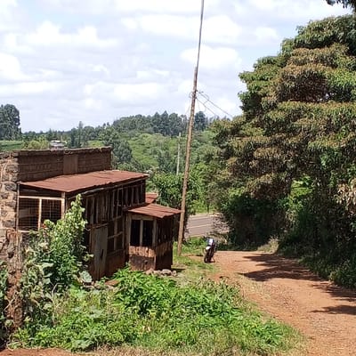 The image depicts a rural scene near Mweiga, Kenya, showing a dilapidated, two-story structure constructed of stone and corrugated metal. Vegetation surrounds the structure, and a dirt road leads past it. A utility pole is nearby, and a motorcycle is parked further down the road, near a paved road in the background. The landscape includes rolling hills covered with trees. The sky is partly cloudy.