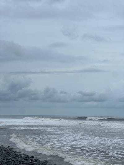 The image depicts a view of the ocean from a pebbled shore in El Tunco, El Salvador. The sky is overcast with heavy gray clouds, suggesting a potentially stormy or cloudy day. The ocean is active with moderate waves breaking and rolling towards the shore, creating white foam. The water appears a muted gray-blue. The foreground consists of a dense scattering of dark, smooth pebbles, indicating a rocky beach. The scene is devoid of any people or specific structures, focusing solely on the natural elements of the coast.