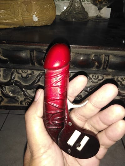 A hand holds a red, phallic-shaped object resembling a derringer pistol. The object has a silver trigger, a dark red handle with two white rectangles and what looks like "PAT. 190689" written on it. The red "barrel" has ridged designs. The hand is positioned against a background of dark furniture with carved designs and a partially visible display of glassware and a light brown carved object. The lighting is dark.