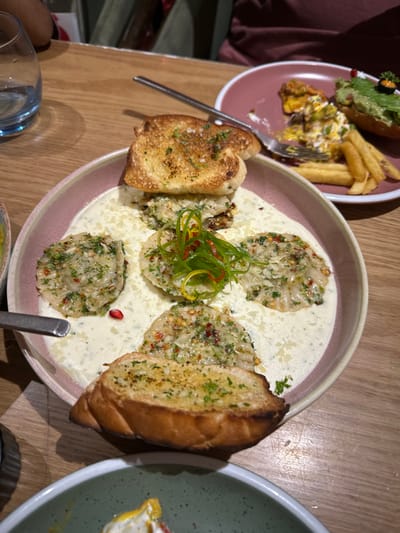 The image displays an indoor restaurant dining scene on a light-colored wooden table. A large, round, pink-rimmed plate centrally features a dish with a creamy white sauce. Within the sauce are five ravioli-like pieces, garnished with chopped green herbs, red chili flakes, and grated white cheese. The central piece includes additional julienned green and yellow strips with red threads. Two toasted garlic bread slices, topped with green herbs, are positioned at the plate's top and bottom. A single red pomegranate seed rests in the sauce.
To the upper right, a smaller, dark pink plate holds partially consumed items: golden-brown french fries, a portion of a yellow and white scramble, and a green avocado toast with a black object and red seed garnish. A silver dinner fork is placed on this plate. A clear, blue-tinted glass of water is visible to the upper left. The handle of a silver utensil is on the main plate's left edge. The rim of a light green, speckled bowl with yellow and white food is partially visible at the bottom.
Blurred figures, one in a reddish-brown garment, are present in the background. The specific city of Kalyan-Dombivli, India, cannot be visually confirmed from the image content.