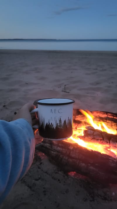 A person's hand, wearing a light blue long-sleeved garment, holds a white enamel mug with a black rim. The mug features a silhouette of pine trees along the bottom and the Cyrillic letters "ЛЕС" (LES) written twice in a lighter color on the upper portion. The mug is positioned in front of a campfire, with bright orange flames and glowing embers visible. The background consists of a sandy beach leading to a calm body of water, with a dusky sky overhead. The scene appears to be set in Zaostrov'e, Russia, likely near the coast, during twilight.