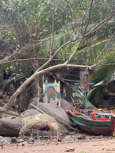A tropical scene featuring lush greenery and a scattering of man-made elements. A leaning tree dominates the foreground, with a backdrop of palm fronds. In the background, there is a hut-like structure adorned with a large painted face and various signs. An old boat sits on the shore nearby. The beach is covered in pebbles, shells, and colorful debris.