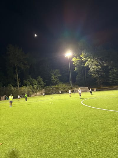 This image depicts an artificial turf sports field at night, located at Albemarle Field, Newton, Massachusetts, United States. The field is illuminated by multiple overhead stadium lights, one of which stands prominently on the right side of the frame, casting bright illumination and a slight lens flare. The sky is dark, with a distinct crescent moon visible in the upper left quadrant.
Seven to nine individuals are scattered across the green artificial turf. On the far left, a person wearing a bright yellow/lime green shirt and blue shorts stands, facing right. Towards the left-center, two individuals are positioned near a small white soccer goal; one wears a light green long-sleeved shirt and dark pants, while the other wears a white shirt and dark shorts, positioned slightly behind. Three white cones or markers are visible on the field in the mid-left. In the mid-ground, a larger white soccer goal is present. Behind this goal and to the right, a white baseball backstop netting is visible, indicating an adjacent baseball diamond. A treeline forms the background perimeter of the field, with a concrete or stone retaining wall visible behind the left soccer goal. Other individuals, dressed in athletic wear, including t-shirts, shorts, and long pants, are observed moving or standing across the field, consistent with a soccer practice or casual game. One person in a grey t-shirt and dark shorts is walking towards the foreground in the mid-right. A prominent white curved line marking is visible on the artificial turf in the foreground on the right.