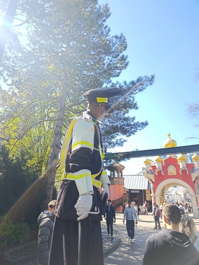 The image shows a tall man on stilts dressed in a black and white ceremonial uniform with yellow accents, wearing a black hat and white gloves, standing on a paved path. He is positioned in front of a crowd of people walking in the same direction. Behind the crowd is a red archway topped with golden domes. The location is in the Europa-Park theme park in Rust, Germany, specifically in the Russian-themed area, evidenced by the architectural style of the archway. A large tree is to the left of the man on stilts. The sky is clear and blue. There is a lens flare from the sun in the upper left of the frame.