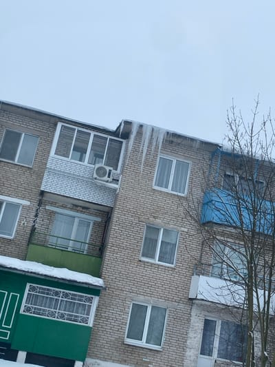 The image depicts a multi-story residential building located in Карабоўскі сельскі Савет, Belarus, under overcast winter conditions. The structure is constructed of light-colored brick with numerous rectangular windows featuring white frames.
Prominently, a series of large, pointed icicles hang from the building's main roof edge, particularly above the left side, indicating freezing temperatures. Below the icicles, on the upper left, is an enclosed loggia with white-framed glass panels and a white brick base, from which an outdoor air conditioning unit is mounted. Directly beneath this, a mid-level balcony features a green metal base and white railings, with snow accumulating on its roof. At the bottom left, another enclosed loggia is visible, predominantly green with white decorative window grilles and snow on its roof.
On the right side of the building, a curved, blue-colored balcony is partially visible, and below it, another open balcony with white railings is obscured by the bare branches of a leafless deciduous tree. The sky is uniformly light gray and overcast. Snow covers visible ground areas and light accumulations are present on external architectural elements. No people or active interactions are depicted in the scene.