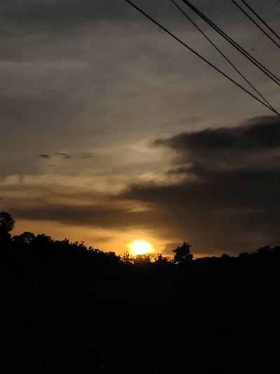 The image shows a dark, overcast sunset in Seuneubok, Indonesia. In the foreground, a silhouette of trees and foliage defines the horizon. The sun, a bright orange orb, is positioned just above the treeline, partially obscured by the branches. The sky is filled with gray and dark clouds, some bearing lighter, yellowish tints from the setting sun. Overhead, electrical wires cut across the upper-right corner of the frame. The light conditions suggest dusk or twilight. There are no people visible in the frame.