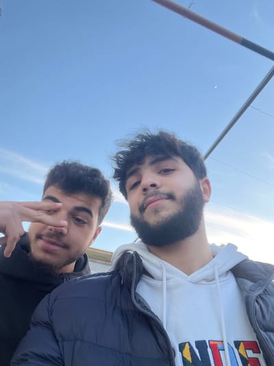 Two young men pose closely for a selfie under a bright, clear sky. The person in the foreground makes a hand gesture, while the other is slightly behind, wearing a white hooded sweatshirt under a dark blue puffer jacket. A metal bar with some wires stretches diagonally across the upper left corner of the frame. The overall image composition tilts slightly.