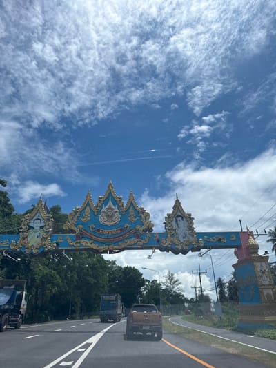 This is a vertical, eye-level shot of an ornate blue and gold archway on a sunny day in Ban Tha Chatchai, Thailand. The archway, which appears to be a gateway to a town or temple, is decorated with images of people and intricate gold scrollwork. Palm trees and other lush green foliage are visible in the background, suggesting a tropical setting. A paved road runs alongside the archway, with a yellow line dividing the lanes. Several vehicles, including a blue truck and a smaller brown truck, are on the road, indicating traffic. The sky is a vibrant blue with scattered white clouds. The time of day appears to be daytime, given the bright sunlight. The overall impression is one of a tropical, possibly religious, landmark in Thailand. Visible text is present on the archway, but it is in a language that cannot be determined from the image.