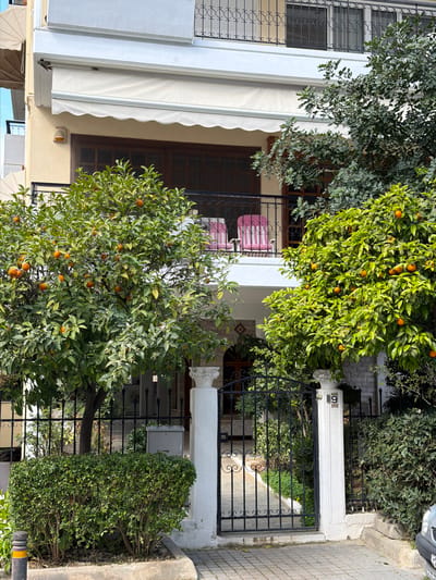 A street-level photographic scene captures the facade of a multi-story residential building located in the Municipal Unit of Elliniko, Greece. The building's lower visible section is finished in a light beige stucco, featuring dark brown wooden-framed windows and balcony doors. On the first floor above ground, an extended off-white retractable awning shades a balcony equipped with a black wrought iron railing showcasing scrollwork. Two pink-and-white striped patio chairs are positioned on this balcony. Above, a second floor displays windows secured with black wrought iron grilles.
The property entrance consists of a black wrought iron gate with decorative elements, flanked by two white, fluted concrete pillars topped with Corinthian-style capitals. The number "9" with "9A" below it is affixed to the right pillar. The ground surface leading to the gate and extending to the right is paved with grey interlocking pavers.
Prominent in the foreground are two large citrus trees laden with ripe orange fruit and white blossoms, positioned on either side of the entrance walkway. To the left of the gate, a dense, trimmed green hedge runs along a black metal fence. Further greenery, including a large tree with dark green foliage, is visible towards the right side of the building and within the property behind the fence. A black bollard with a yellow reflective stripe is visible on the far left at the curb. The front end of a silver passenger vehicle is partially visible, parked on the street on the far right. No individuals are present in the image.