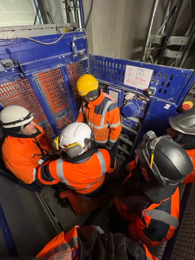 Inside a personnel/material hoist at a construction site in Aulnay-sous-Bois, France, five male construction workers are gathered. The hoist structure is primarily blue metal with grated panels. A warning sticker on the interior wall indicates a "CMU : 1500 Kg" (Maximum Useful Load: 1500 kg) and displays safety pictograms. One worker, centrally positioned and facing inward, wears a yellow hard hat and an orange high-visibility jacket with grey accents. To his left, another worker with a white hard hat and headlamp wears a similar orange jacket, looking towards the center. Below him, a third worker in a white hard hat with ear protection and an orange jacket also looks towards the center. Two additional workers, partially visible on the right and bottom right, wear black and metallic grey hard hats, respectively, along with orange high-visibility jackets. All individuals are dressed in work attire consistent with construction environments. The workers are in close proximity, appearing to be in discussion or inspecting the hoist's internal components, which include machinery elements and a control panel with red and yellow buttons visible on the right. The hoist shaft's concrete walls and metal guide rails are visible in the background.