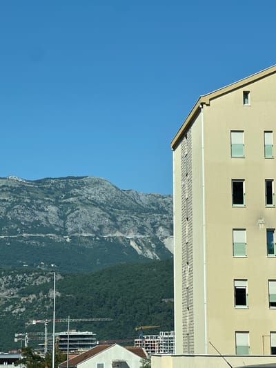 The image depicts a sunny day in Prijeradi, Montenegro, with a clear blue sky dominating the upper portion of the frame. In the foreground, on the right side of the image, is a light yellow building with several windows. Some windows have white blinds partially lowered, while others are dark, indicating they are closed. The building has a textured vertical band running down its side.

In the middle ground, a range of green, tree-covered hills slopes upwards towards rocky, grey mountains in the background. A road or path is visible winding along the mountainside.

Further in the middle ground, below the hills and to the left of the yellow building, a construction site is visible with several tall cranes and partially built structures. A street light stands in front of the construction site.

In the lower foreground, the edge of another building with a terracotta-tiled roof is partially visible. The overall scene suggests a developing urban area with a backdrop of natural mountainous landscape. No people are visible in the image.