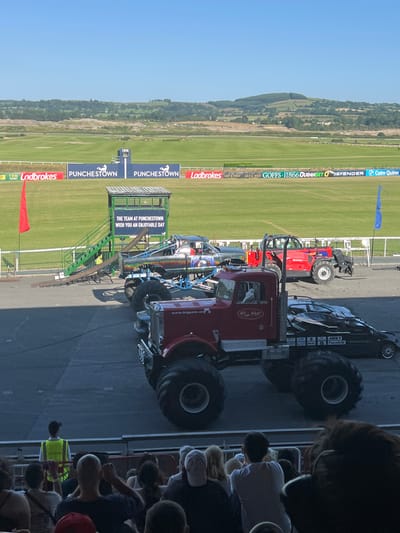 This image captures a sunny day at Punchestown Racecourse in Ireland, with a clear blue sky. In the foreground, a large red monster truck is positioned on a paved area. To its right, a black car is parked. Behind these vehicles, a red telehandler is visible. Further back, a monster truck with a colorful design is on a ramp. A green structure with stairs stands next to the ramp. In the distance, a lush green racecourse stretches out, with rolling hills and trees forming the backdrop. Banners displaying sponsor logos are visible along the edge of the racecourse. A few people are seen in the foreground, some appearing to be spectators. A person in a high-visibility vest is also present near the ramp. The overall scene suggests an outdoor event with vehicles on display.
