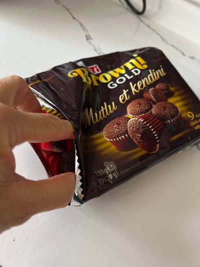 A hand is opening a package of "Browni Gold" chocolate cakes. The package has a dark brown background with yellow lettering. The image of the cakes shows several small, chocolate-covered cupcakes. The package is resting on a white, speckled surface, likely a countertop. The scene is indoors, with a blurred background that suggests an indoor environment. There is no indication of the city of Ümraniye, Turkey, within the image.