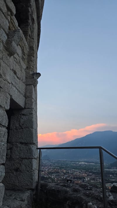 The image depicts an outdoor scene from a high vantage point, likely a historic ruin or fortification, looking out over a valley and a town at dusk. In the foreground, massive, weathered stone blocks form a wall or walkway, suggesting ancient architecture. A small, dark object, possibly a bird, is perched on the stonework.

To the right of the stone structures, a modern metal railing with multiple horizontal wires creates a barrier, separating the viewer from the precipice. Through the railing, a panoramic view of a town unfolds. Buildings with terracotta roofs are nestled among green fields, and winding roads can be seen. In the distance, dark, imposing mountains rise, their slopes partially obscured by the fading light.

The sky is a dominant feature, transitioning from a pale blue at the top to warmer hues near the horizon. A vibrant band of orange and pink clouds stretches across the sky, catching the last rays of the setting sun. This colorful cloud formation is a prominent detail, indicating the time of day is sunset. The overall atmosphere is serene and picturesque, evoking a sense of history and natural beauty. The location context suggests this is Riva del Garda, Italy, a town known for its mountainous backdrop and proximity to Lake Garda. There is no visible text in the image.