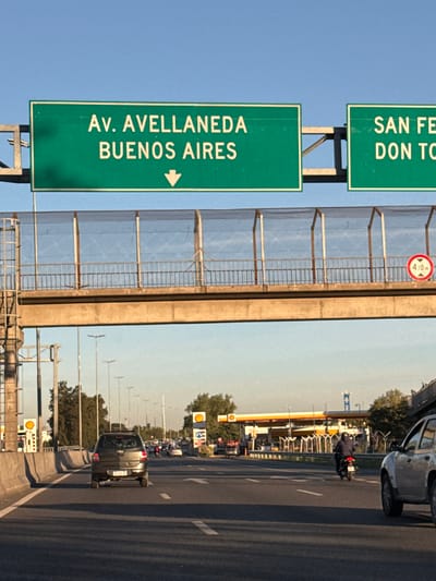 The image shows a road scene in San Fernando, Argentina. The road has multiple lanes and appears to be a highway. There are vehicles on the road, including cars and a motorcycle.

Road signs are visible, indicating directions to "Av. Avellaneda Buenos Aires" and "San Fernando/Don Torcuato". There is also a sign indicating a height restriction of 4.10 meters.

A Shell gas station is seen on the side of the road. The weather appears to be sunny. There are trees and streetlights along the roadside.