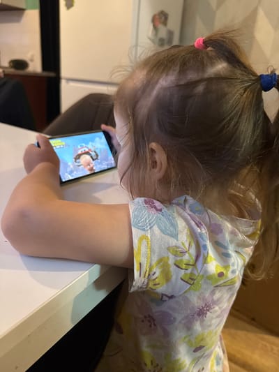 A close-up, side-profile shot shows a young child with brown hair tied into two pigtails, one with a pink hair tie and the other with a blue one. The child wears a light-colored shirt with a floral pattern of purples, yellows, and blues. They are looking down at a smartphone held in their hands, which displays a cartoon image of a character in an aviator outfit. The child's arm is resting on a white surface, possibly a table, and the background is blurry but suggests an indoor setting with hints of furniture and possibly shelves. The lighting appears to be artificial. No specific time of day or weather conditions are discernible. The child's expression is focused on the screen. There is no visible text in the image.