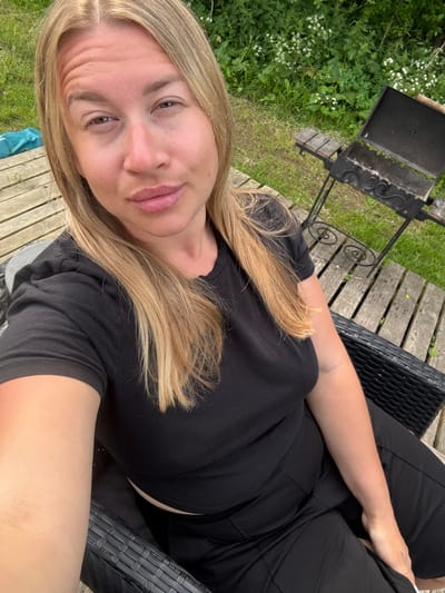 A blonde woman with fair skin and pink lipstick is sitting on a dark wicker chair outdoors. She is wearing a black t-shirt and black pants. Her hair is shoulder-length and blonde. Behind her, a wooden deck is visible, with a grill and green foliage in the background. The scene appears to be a backyard in Zamosh'e (Zaklinskoe S/P), Russia.