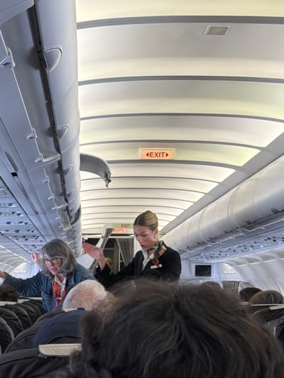 The image captures the interior of an airplane cabin, likely an aisle view looking towards the front or rear of the aircraft, presented at a tilted angle. The scene is brightly lit by artificial cabin lighting.
In the mid-ground, a flight attendant with blonde hair, wearing a dark uniform jacket and a patterned scarf, stands in the aisle, facing right and appearing to gesture with her hands, possibly directing or assisting passengers. Behind her, two prominent red "EXIT" signs with directional arrows are visible on the overhead panels.
To the right of the flight attendant, an older woman with gray hair and glasses, dressed in a denim jacket and a patterned scarf, is seen bending slightly in the aisle. Partially visible in front of her is an older man with white hair and a dark blue cap. The back of a person's head with dark, possibly furry, hair occupies a large portion of the foreground on the right, suggesting the perspective is from behind a passenger.
The left side of the frame showcases the airplane's interior architecture, with closed overhead bins and curved, light-colored panels along the ceiling and walls, characteristic of an aircraft. Individual reading lights and air vents are visible on the underside of the overhead compartments. Faint labels like "A B C" are discernible on the side of seats or storage compartments below the bins.
The activity suggests passengers are either boarding, deplaning, or moving through the cabin. Given the location context, this scene takes place inside an aircraft at Kloten, Switzerland (Zurich Airport). The overall atmosphere appears calm.