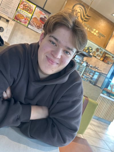 An individual is depicted seated at a light-colored table inside a Coffee Bean & Tea Leaf establishment in Medford, United States. The subject is a young person, appearing male, with light brown hair parted in the middle and blue-green eyes. They wear a dark brown hooded sweatshirt and are looking directly at the camera with a closed-lip smile, arms crossed over the chest. The background reveals two vertical menu boards displaying food items such as sandwiches and soups, and a glass display case containing various baked goods and pastries. A prominent wooden sign bearing the "THE COFFEE BEAN & TEA LEAF" logo and text is affixed to a wall behind the display case. Seating with a lime green backrest and a light brown cushion, along with a tiled floor, are also partially visible in the brightly lit interior.