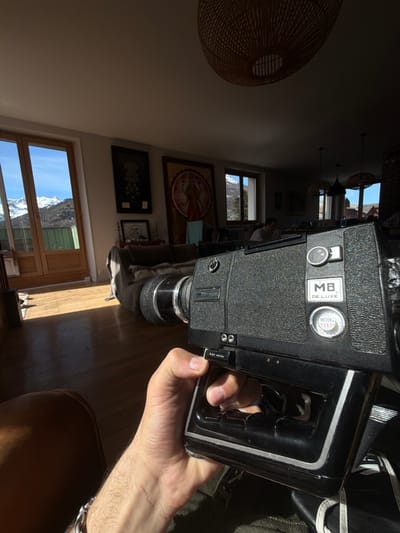 The image captures an indoor residential scene in Briançon, France, featuring a hand holding a vintage film camera with a bright mountainous backdrop visible through windows. In the foreground, a light-skinned right hand with visible arm hair and wearing a silver-colored chain bracelet firmly grasps a black Super 8 film camera. The camera body is textured, bears the labels "MB DE LUXE" and "METER FEET," and has an "OPTICAL-ZOOM" lens protruding from the front. A "SLOW MOTION" toggle switch is present on the camera's grip.

The camera points into a sunlit living area with a light brown hardwood floor. Strong sunlight streams through large double wooden doors on the left and a window on the right, casting bright reflections on the floor. Through these openings, a clear blue sky and prominent snow-capped mountains are visible in the distance, with patches of green vegetation closer to the structure. Distant buildings with reddish roofs are discernible through the far-right window.

Inside the room, a dark-colored sofa is positioned in the mid-ground. Above it, two large framed artworks are mounted on a light-colored wall: one dark and abstract, the other a vibrant, circular mandala-like painting with a central figure. A dark, woven pendant light fixture hangs from the ceiling. Further in the background, a person is seated at a table, facing away from the camera, partially obscured by shadows.