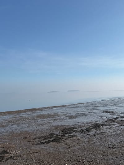 The image displays a coastal scene, likely from the foreshore of Penarth, United Kingdom, looking out over the Bristol Channel. The sky, occupying the upper two-thirds, is clear and light blue, with a subtle atmospheric haze merging with the distant horizon. In the middle ground, two distinct, dark landmasses, consistent with Flat Holm and Steep Holm islands, are visible, partially obscured by the haze over the calm, light blue-grey water. The foreground reveals an exposed, wet foreshore composed of dark brown mud, lighter grey-brown pebbles, and scattered patches of dark, possibly organic, material. The ground glistens in areas, indicating dampness or recent tidal exposure. No individuals, structures, or actions are depicted.