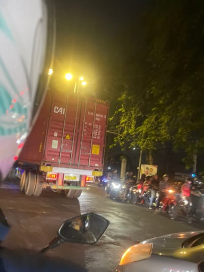 The image depicts a nighttime scene at an intersection in Sruni, Indonesia. A large red shipping container truck, identified by the "CAI" logo, is the dominant subject, positioned on the left side of the frame. The truck displays a serial number "464762 0 4561" and a "CAUTION 9'6" HIGH" warning sticker. Its rear lights are illuminated, and the wheels are visible.
To the right of the truck, a line of motorcycles with riders, many wearing helmets, are stopped, suggesting a traffic jam. The motorcycles have their headlights and taillights on. The road surface appears wet, reflecting the lights. In the background, trees with illuminated foliage are visible, along with streetlights. A poster with graphics is also present, partially obscured by the trees and motorcycles. The overall impression is a busy urban intersection at night, likely experiencing traffic congestion. The foreground shows a partial view of a motorcycle's side mirror and a car's headlight, indicating the viewer is likely on a motorcycle or in a vehicle.