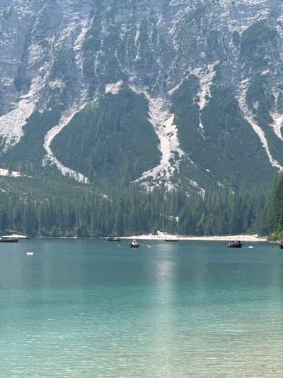 The image depicts a serene mountain lake, likely Pragser Wildsee in Prags - Braies, Italy. In the foreground, the water exhibits a striking gradient of turquoise and clear blue hues, with subtle ripples on its surface. Several small wooden rowboats are scattered across the lake, some with occupants rowing. In the mid-ground, a sandy shoreline is visible, dotted with numerous people enjoying the scenery. Behind the shoreline, a dense forest of evergreen trees stretches upwards, meeting the imposing, rugged slopes of a large mountain. The mountain face itself is characterized by steep, rocky terrain interspersed with patches of green vegetation, and a prominent white scar running diagonally down its side. The sky is not visible in the frame.