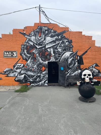 The image depicts the exterior of a building in Voskhod, Russia, featuring a large, intricate graffiti mural on an orange brick wall. The mural is a detailed black, white, and orange depiction of a stylized skull with robotic and mechanical elements, dominating the wall. To the left of the entrance, a sign reads "ЗАЛ № 3 МУЗЕЯ" (Hall No. 3 of the Museum).

In front of the mural and to the right of an open, dark doorway, stands a large, black, rounded sculpture of the Grim Reaper, complete with a skull face, black hood, and holding a scythe with a skull flag. A metal bench is situated near the doorway, partially obscured by the sculpture. The ground in front of the building is paved with asphalt, showing some wear and cracks, with a small patch of green grass growing to the left of the doorway. Overhead, electrical wires are strung across the sky. The sky is overcast and a pale gray. The overall scene suggests an industrial or artistic location, possibly an entrance to a museum or gallery with a distinctive, dark aesthetic.