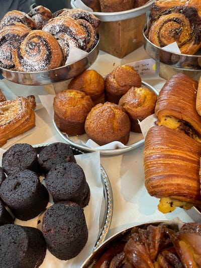A bakery display features an assortment of pastries in silver trays lined with white paper. On the left, trays hold chocolate swirls dusted with powdered sugar. In the center, golden muffins are nestled together. To the right, cylinders of dark, almost burnt, cake or bread. Top trays are filled with croissants and other laminated dough pastries. The scene presents a tempting array of baked goods.