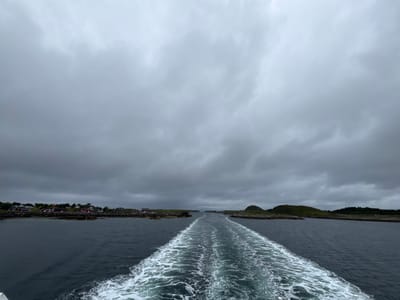 The image depicts a view from a boat moving away from a settlement on Sleneset, Norway. The water is dark and choppy, with a prominent white wake trailing behind the boat. The shore, visible on the left, is lined with red-roofed buildings and green, rocky terrain. The sky is overcast with heavy grey clouds. The scene suggests a journey through a fjord or coastal area under a cloudy, possibly cool, day. There are no people visible in the image.