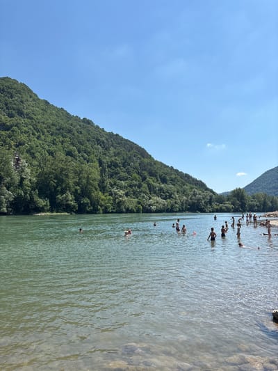 A wide, clear river flows through a lush, green valley under a bright blue sky with a few wispy clouds. The riverbanks are lined with dense trees, and steep, tree-covered hills rise on either side. Numerous people, including adults and children, are bathing and playing in the shallow, clear water near the shore. Some are wading, while others are swimming or standing in the water. The water is calm, with gentle ripples reflecting the sunlight. The scene appears to be a popular spot for recreation and enjoying the natural beauty of the area. This location is the Drina river in Zvornik, Bosnia and Herzegovina.