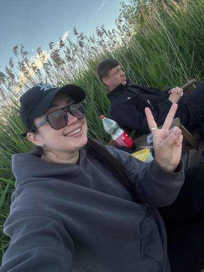A medium shot shows two people relaxing outdoors in Khryashchevka, Russia. Tall reeds and blue sky are visible in the background. The person closest to the camera is wearing a black baseball cap, gray hooded sweatshirt, and sunglasses. The person’s right hand is held up in a peace sign. Behind and to the left of the first person, a second person is wearing a black jacket and is seated on a wooden bench. A plastic bottle and container are between the two people.