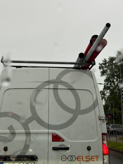 A white utility van is parked outdoors in what appears to be an urban environment. The van is facing away from the camera, displaying its rear doors. On the roof of the van, a metal rack holds several pipes, some of which are red and others appear to be metal. The van is decorated with a large, grey circular logo that resembles a stylized infinity symbol or intertwined rings. Below the logo, on the right side of the rear door, the word "ELSET" is visible in orange lettering, accompanied by a symbol that looks like a power plug intertwined with two circles. The van's license plate is partially visible, indicating a Latvian registration. The scene suggests a cloudy or overcast day, with raindrops visible on the camera lens or the van's surface, indicating recent or ongoing precipitation. In the background, trees and a traffic light are discernible, with a dark-colored car to the right. The image is likely taken in the vicinity of Ķekava, Latvia, as suggested by the license plate and the overall context.