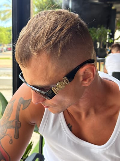 This is a close-up, rotated, vertical photograph of a person's head and shoulder, presumably outdoors in La Vila Joiosa, Spain. The subject is wearing a white tank top and large, fashionable sunglasses with black frames and a gold emblem on the temple. The sunglasses are positioned on their nose and the side of their face, obscuring the eye. The person has short, light brown hair and visible stubble on their chin and neck. There is a tattoo of an anchor on their left arm. In the background, blurred greenery suggests a lush, outdoor environment. The lighting indicates it is daytime, likely sunny. The overall impression is casual and stylish, with the focus on the person's accessories and facial profile.