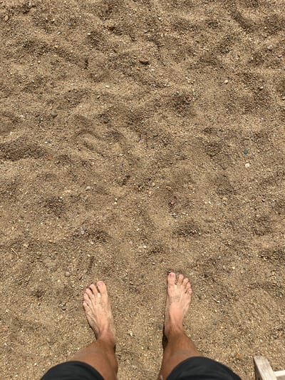 In Gres-2, Kazakhstan, a close-up overhead shot reveals a person's bare feet standing on a sandy surface composed of fine grains and small pebbles. The sand is a light brown color, with some areas appearing slightly darker and damp, indicating a recent presence of moisture or shadow. The feet, dusted with sand, are positioned towards the bottom of the frame, with the toes spread slightly on the ground. The person is wearing black shorts, visible at the very bottom of the image. The surrounding environment is entirely sand, with subtle variations in texture and color, suggesting an outdoor, possibly beach or desert-like setting.