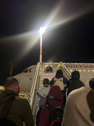 The image depicts a nighttime scene at Emelyanovo Airport, Russia, focusing on passengers boarding a commercial aircraft. A line of approximately seven to ten individuals, mostly viewed from behind, is ascending mobile boarding stairs to the aircraft's front-left entry door. The aircraft is a white narrow-body jet, featuring "РОССИЯ" (Russia) in red capital letters on its fuselage and "Саратов" (Saratov) below the cockpit windows. A bright, tall airport light pole with red and white striped sections stands to the left of the stairs, emitting a strong white glare that dominates the upper central portion of the frame. One person is partially visible entering the aircraft door, while others follow in succession. Distant airport infrastructure or a control tower with red and white markings is faintly visible in the far left background against the dark night sky. The primary light sources are the airport pole, the aircraft's interior, and external boarding area lighting.