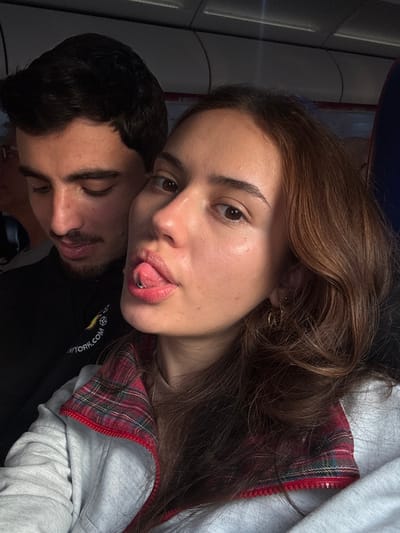 The image captures a close-up, selfie-style portrait of two young adults inside an enclosed cabin environment. The specific location within Yehud Monosson, Israel, cannot be identified from the visual data.

In the foreground and to the right, a female with light brown, wavy hair looks directly at the camera. She extends her tongue, revealing a silver-colored barbell piercing. She wears small gold hoop earrings in her visible right ear and a light-colored, possibly grey, zip-up jacket with a red and green plaid collar, over a tan-colored top. Her complexion is fair, and her eyes are brown.

Behind her and to the left, a male with dark hair, a light mustache, and a beard is visible. He is looking downwards and to his right, not engaging with the camera. He wears a dark-colored top, which displays the white text "NEW YORK.com" and a small yellow graphic on the left chest area.

The background shows the interior of a cabin, characterized by curved overhead panels, possibly white or light grey, and faint red lines along the side, suggesting a mode of transport such as a train, bus, or aircraft. Overhead lighting fixtures are partially visible. The two individuals are seated in close proximity.