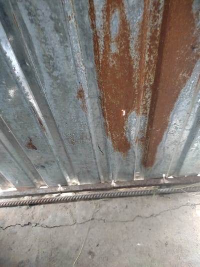 The image shows a close-up of a corrugated metal sheet, likely a part of a fence or shed, in Kegeti, Kyrgyzstan, at the coordinates provided. The metal sheet is rusted in patches, with the rust concentrated on the right side, indicating potential exposure to water or weather. The metal is also showing signs of wear and tear.  At the bottom, a rusted, flat steel bar is secured to the metal sheet, which is attached to a concrete surface. The concrete surface is cracked and shows signs of aging. There are no people or other subjects present.  The image suggests a simple, utilitarian scene, common in urban or semi-urban areas.